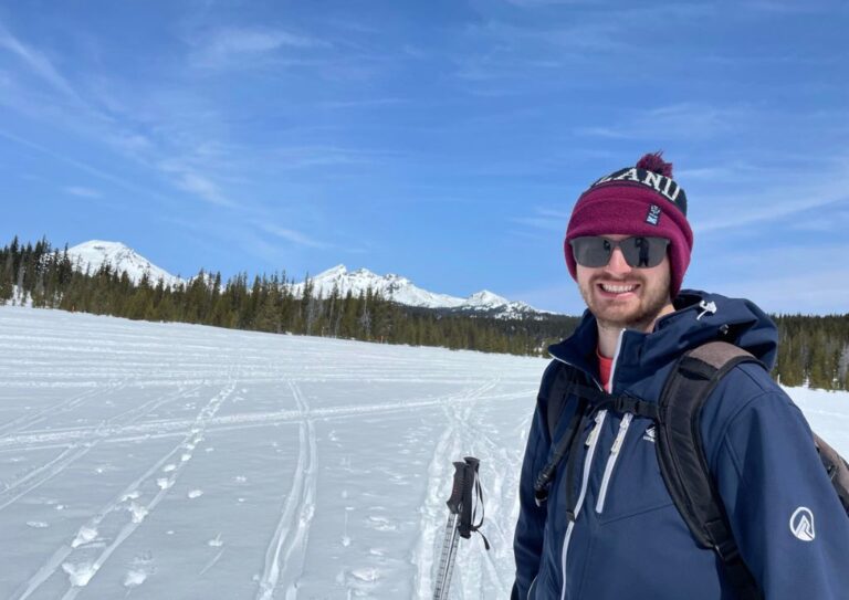 Tyler standing in a snow covered field wearing a jacket and hat to stay warm while hiking.